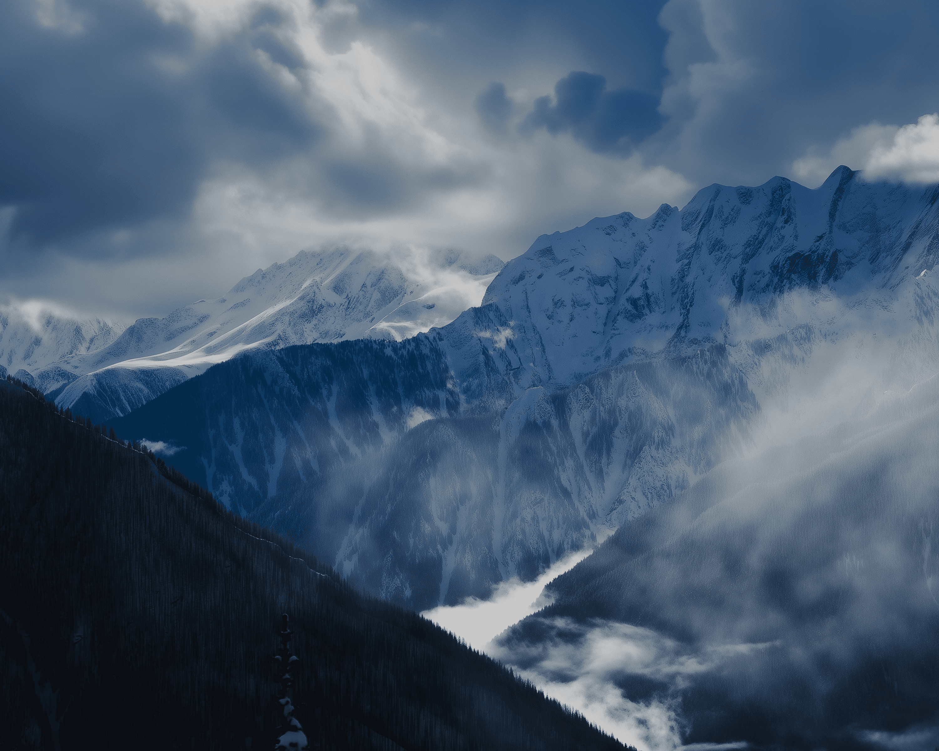 Snowy, jagged mountain range with a blue tint under a dramatic cloudy sky; mist coils through valleys and dark forested slopes rise in the foreground.