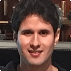 Portrait of a person with short dark hair and a friendly smile, wearing a dark shirt, indoors with a blurred, warm-toned background.