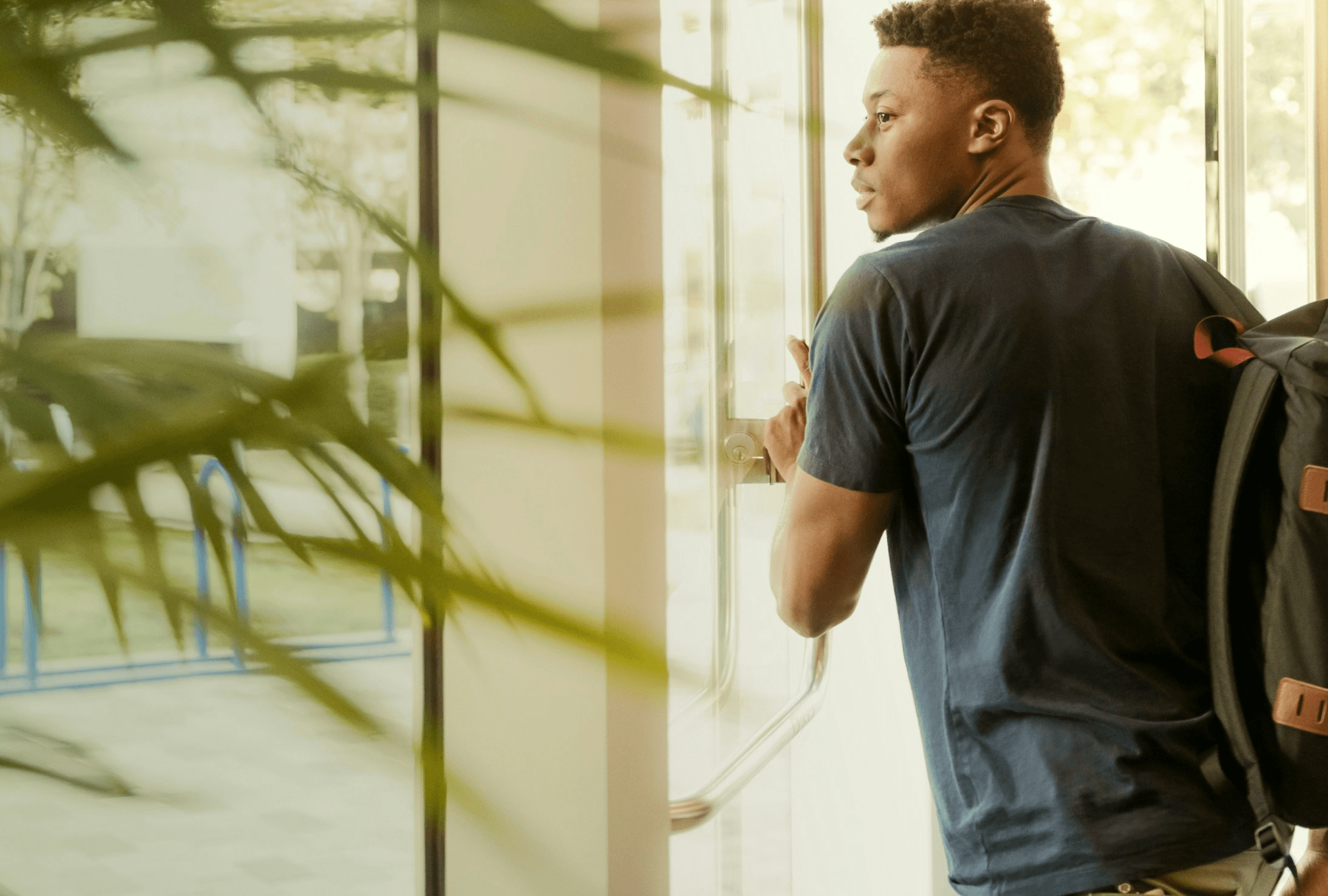 Person with short curly hair, wearing a dark T‑shirt and backpack, stands by a glass door looking outside; a foreground plant partially obscures the view.