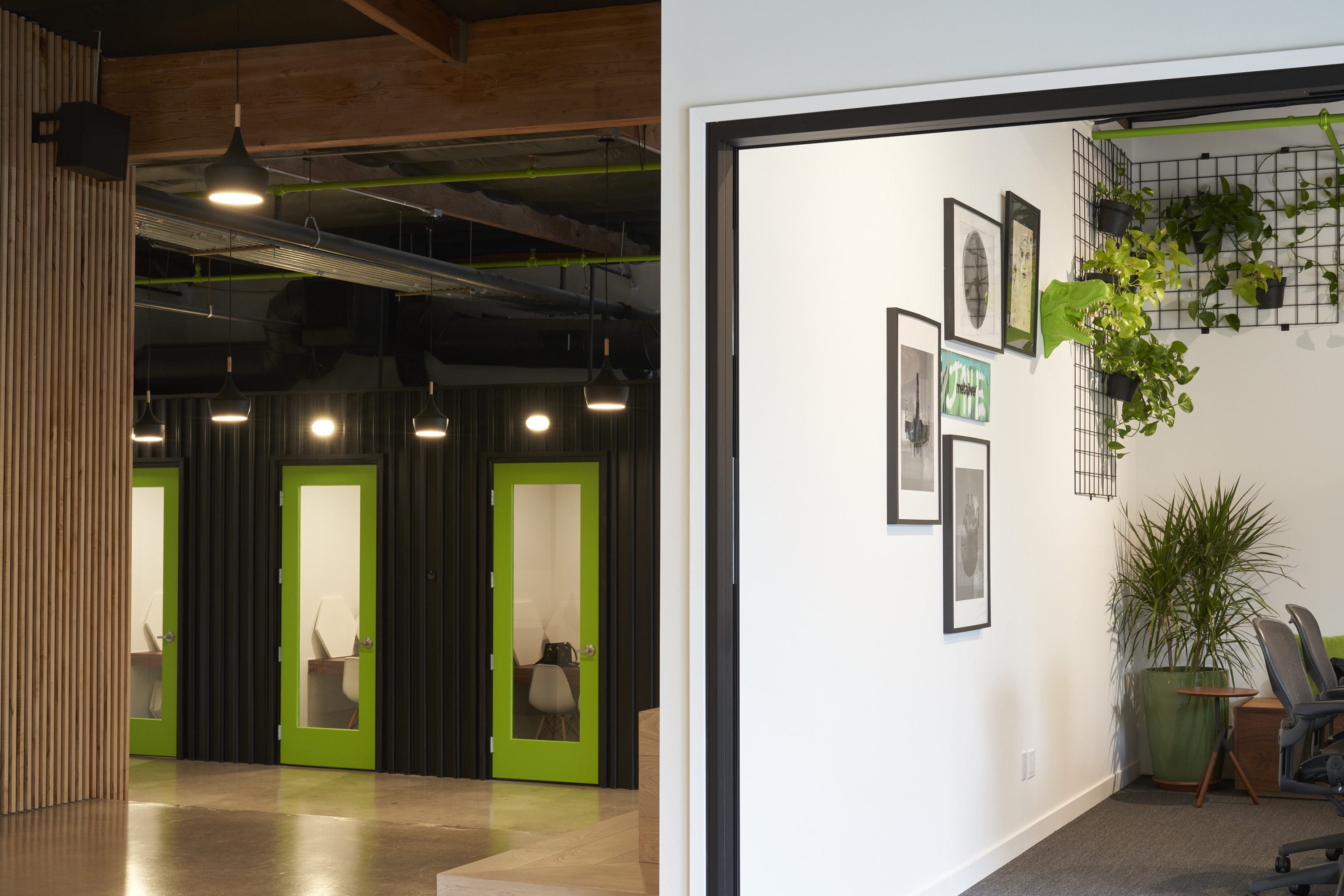 Office interior with lime-green framed glass doors on a dark wall, exposed ceiling pipes and pendant lights; right wall features framed art and hanging plants.