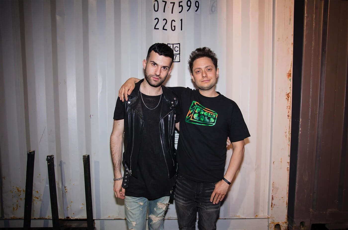 Two people with arms around each other stand in front of a rusted container wall. One wears a black leather vest, the other a black graphic tee.