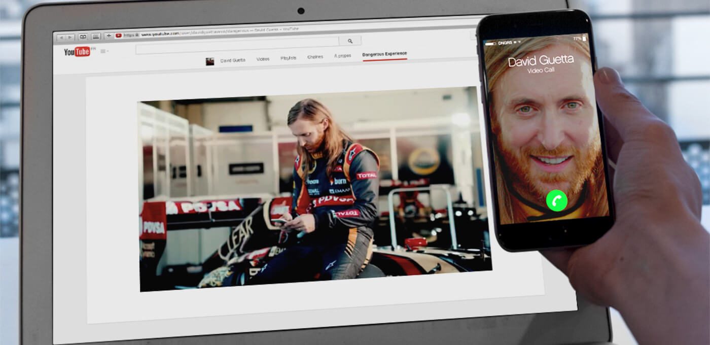 A laptop screen shows YouTube with a racer in a garage beside a race car; a hand holds a smartphone displaying a video call with a bearded person.