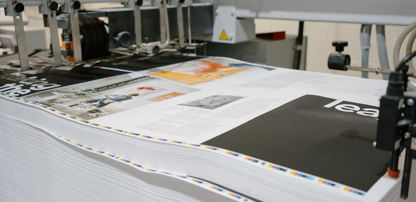 Printing press with stacked pages and color bars along the edge, industrial machinery in the background.