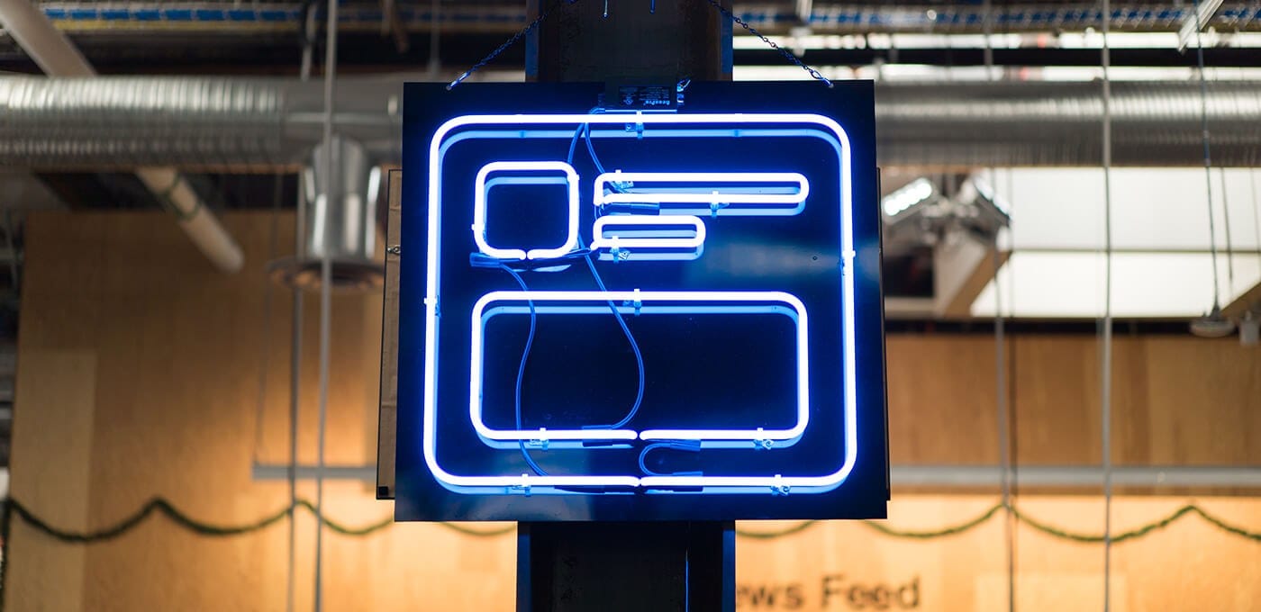 Blue neon sign with a rounded-square outline and stacked rectangles, hanging from the ceiling in industrial space with exposed ducts and wood panels.