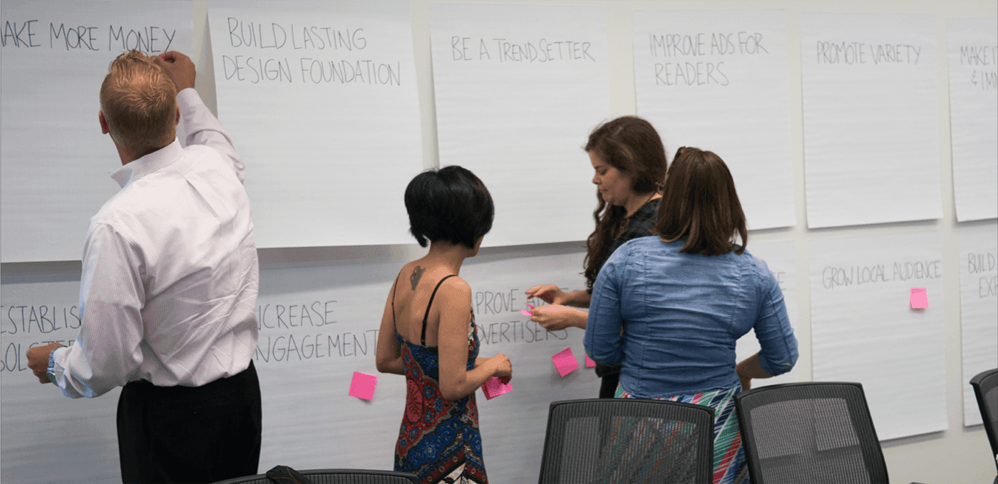Group collaborating in a workshop, adding pink sticky notes to large flipchart sheets bearing business phrases in a bright conference room.