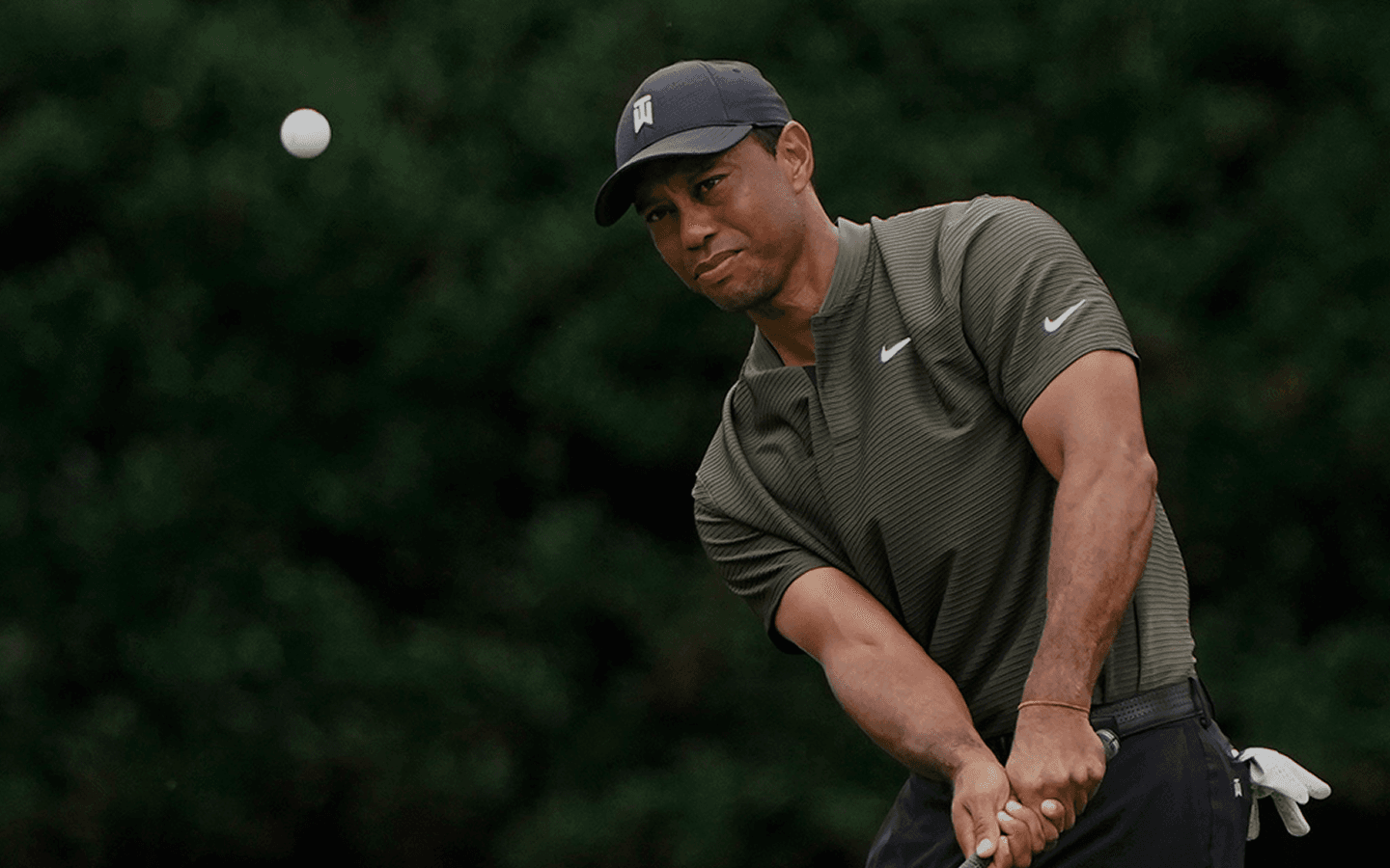 A close-up action shot of professional golfer Tiger Woods wearing a dark cap and polo shirt, swinging a golf club as the ball soars through the air against a blurred, wooded background.