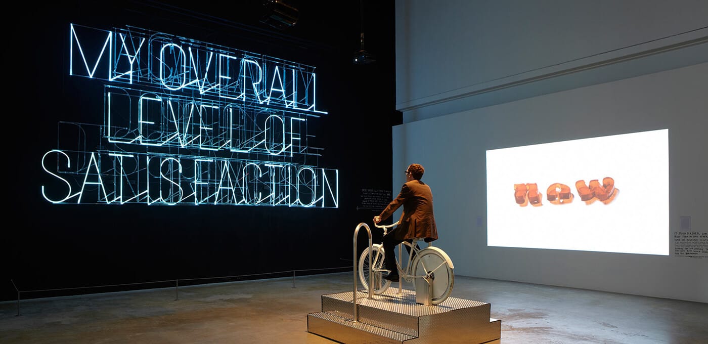 Person rides a stationary bicycle sculpture on a raised metal platform in a gallery; blue neon text glows on a black wall, while a white wall shows a projection of orange letters.