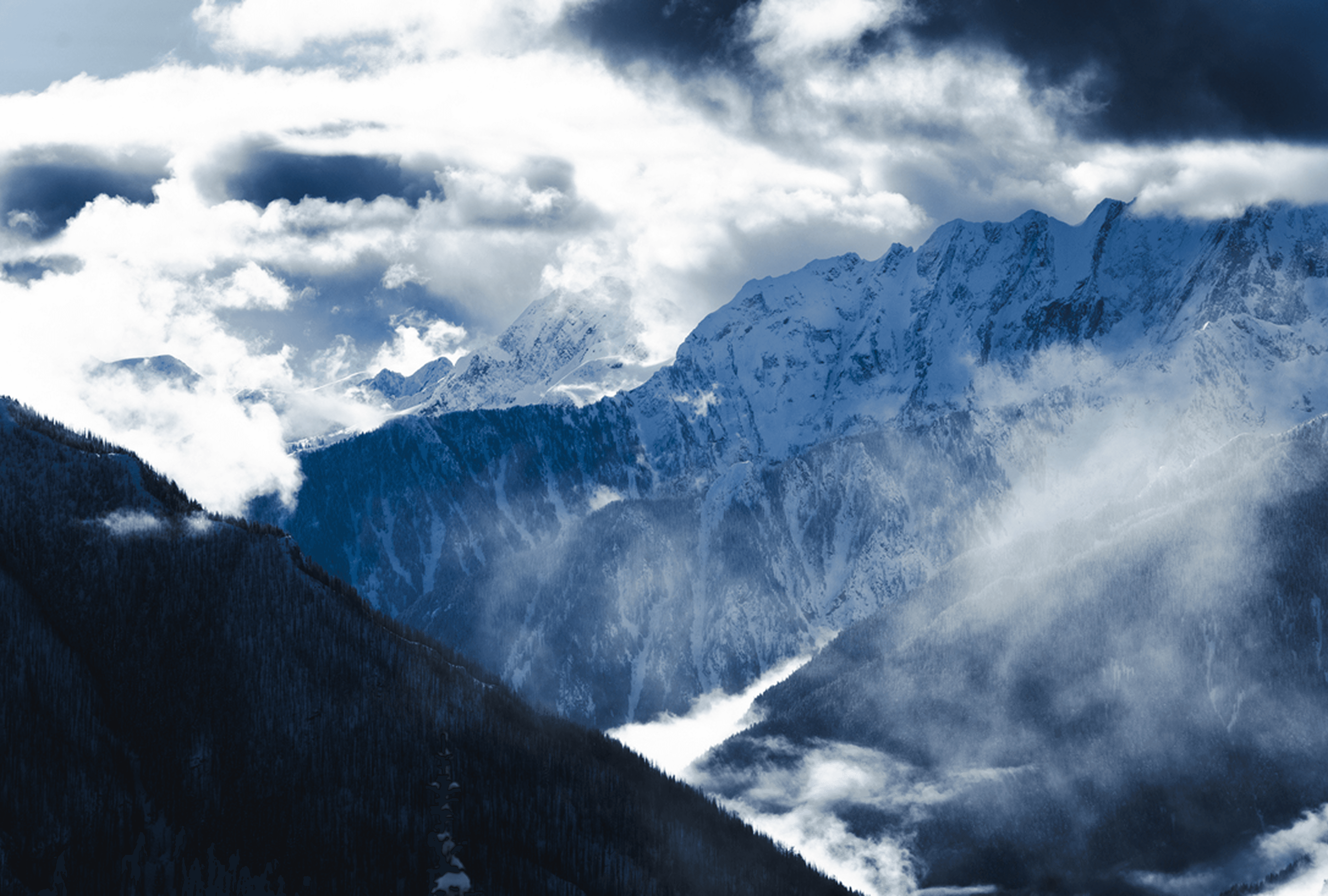 Snow-covered mountain range with jagged peaks, dark forested slopes, and wispy clouds weaving through the valleys.