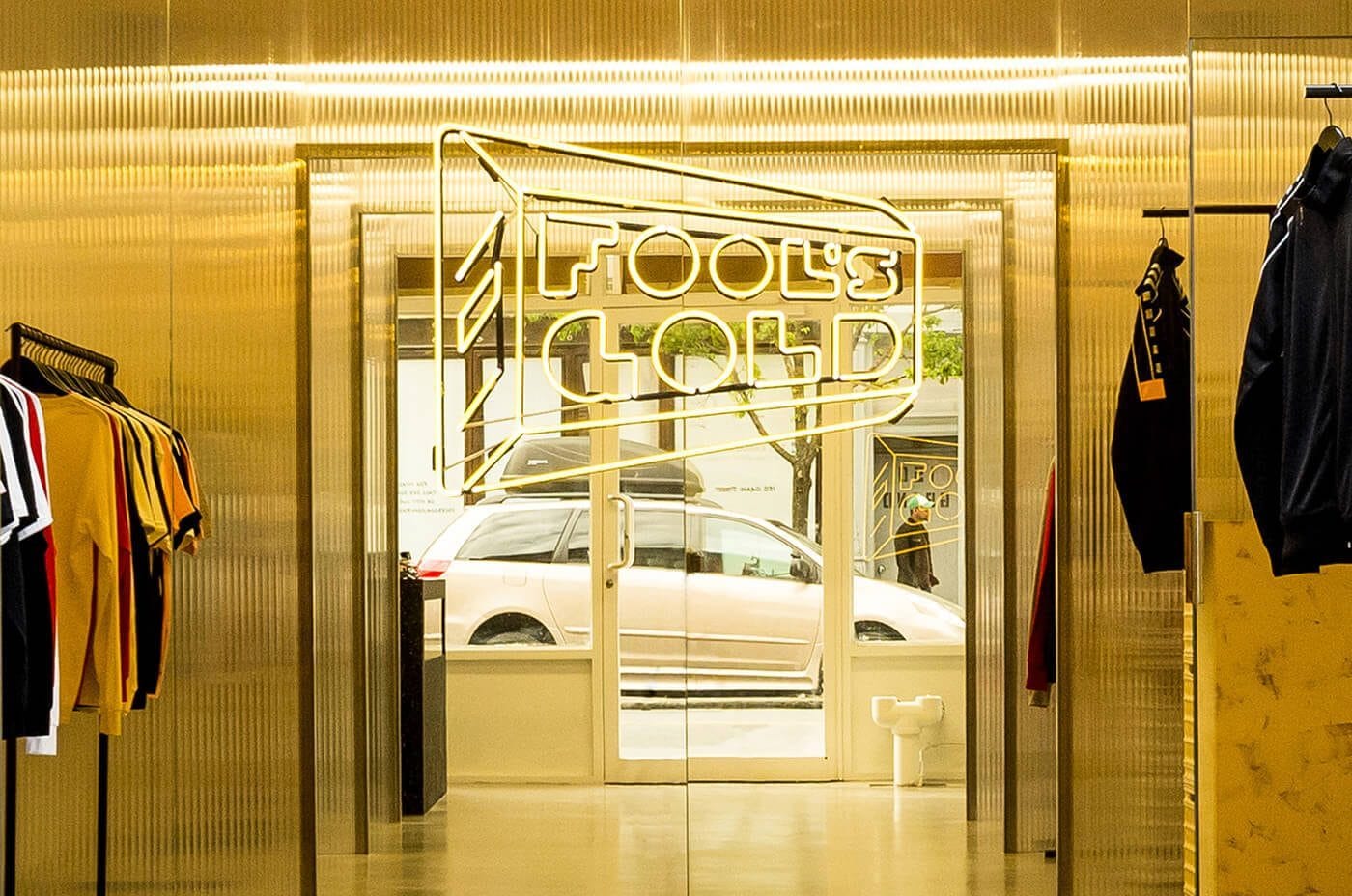 Gold-toned boutique interior with racks of shirts on both sides; neon sign reading FOOL'S GOLD above glass doors, street scene and a car outside.