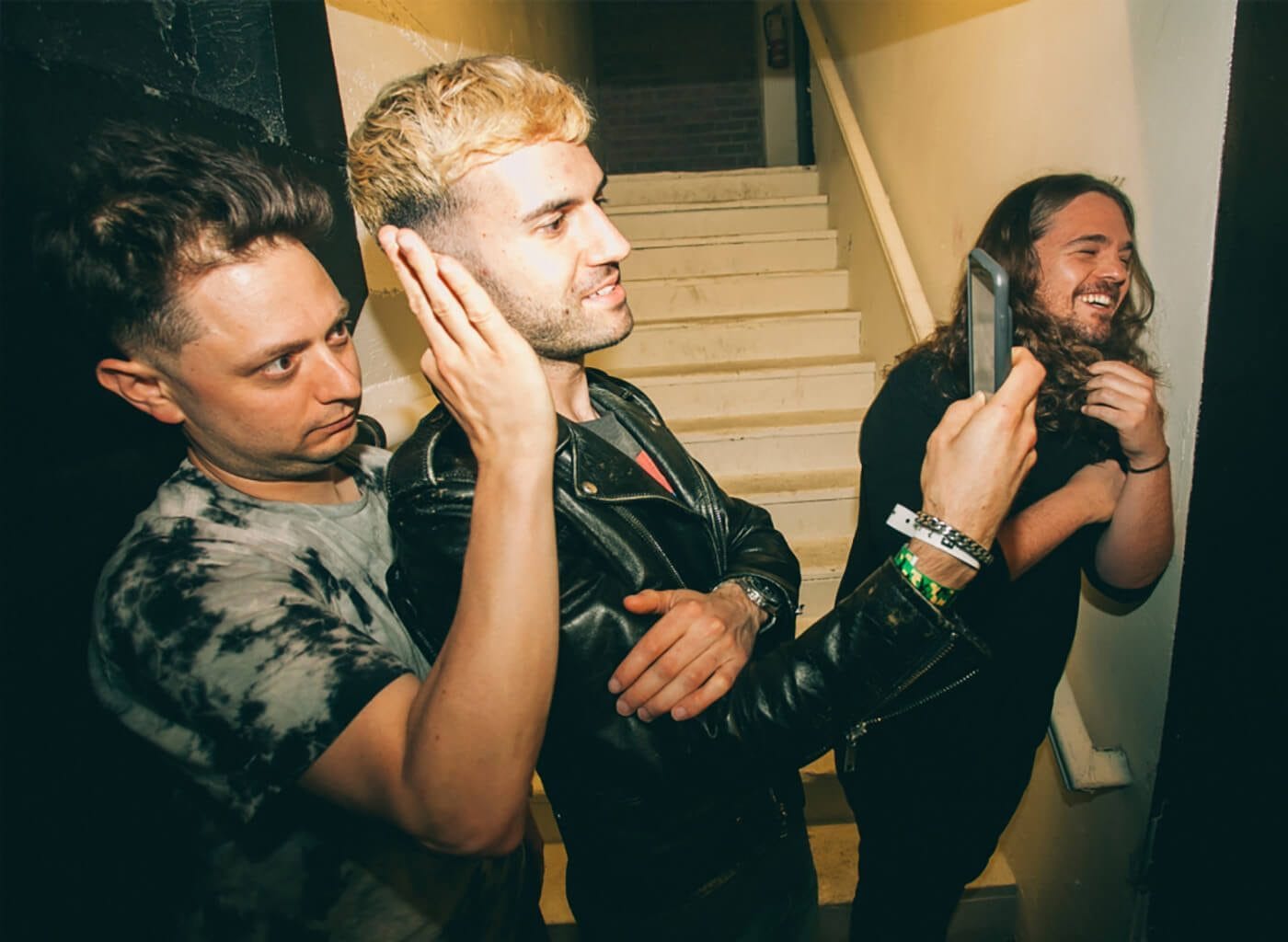 Three people in a stairwell: a blonde person in a leather jacket listens with a hand to the ear, a tie-dye shirted person stands close, and a long-haired person laughs while holding a tablet.