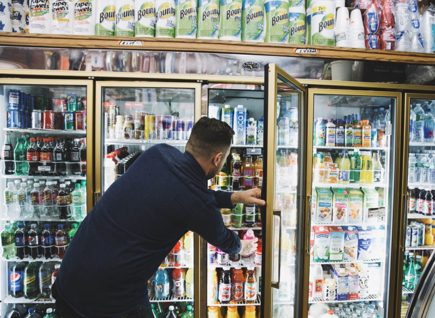 Person in a dark blue sweater reaches into a glass-door refrigerated display, grabbing a pink drink among colorful bottled beverages in a store.