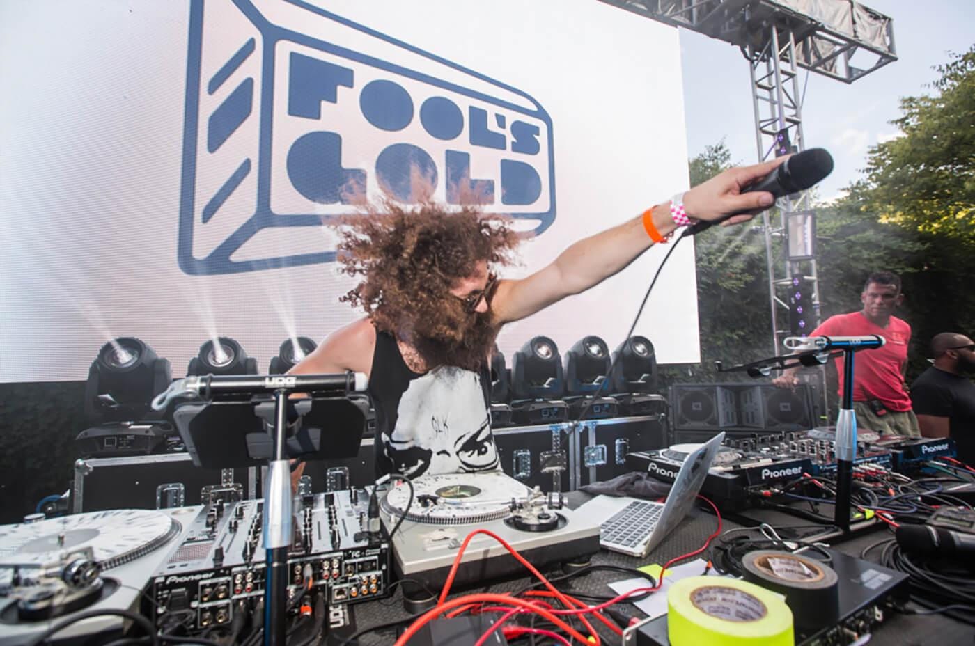 A DJ with long curly hair and a beard performs outdoors, reaching out with a mic over a table of turntables, mixer, laptop, and cables.