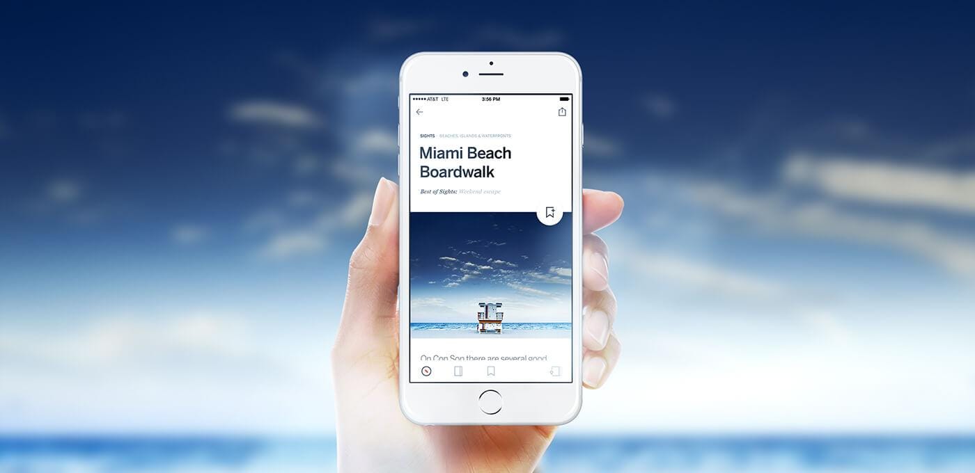 Hand holding a white smartphone displaying a beach travel app with a photo of a lifeguard chair on the sand and blue sky and ocean.