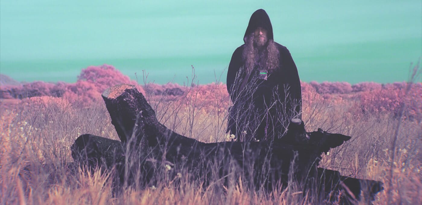 Hooded figure with a long beard stands in tall grass beside a large dark animal carcass, under a turquoise sky with pink-tinted shrubs in the distance.