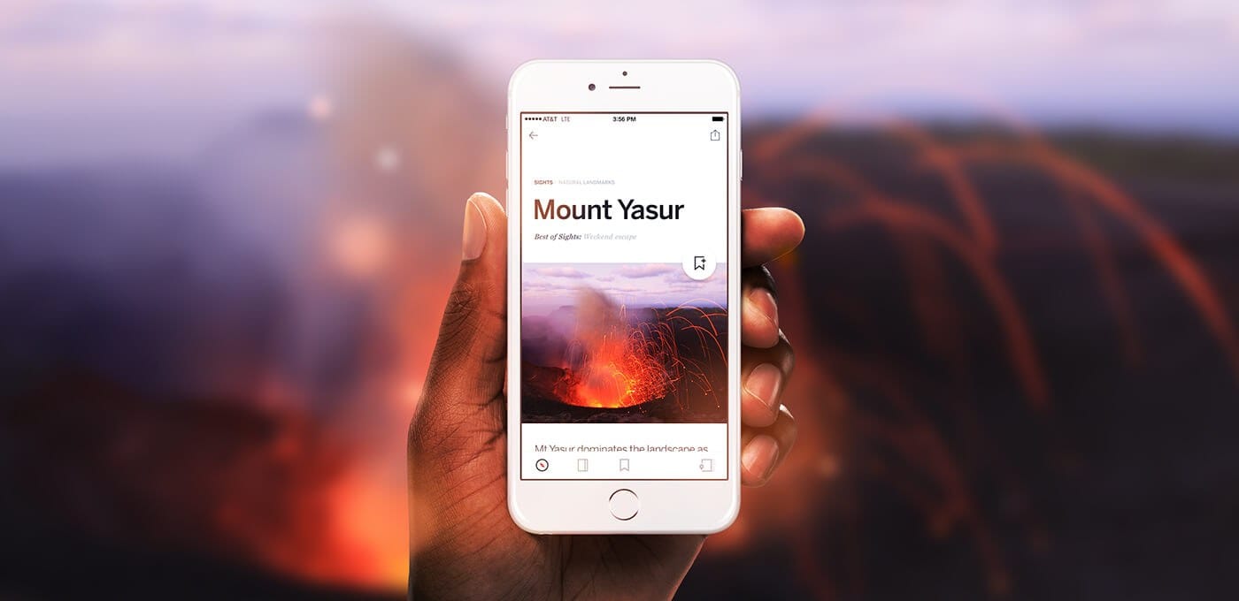 Hand holding a white smartphone displaying a page titled "Mount Yasur" with a volcanic image; blurred lava-lit landscape in the background.