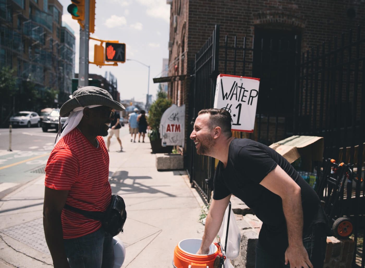 Two people on a sunlit city sidewalk share a smile; one wearing a red striped shirt and hat, the other in a black shirt leaning forward near a "Water $1" sign.