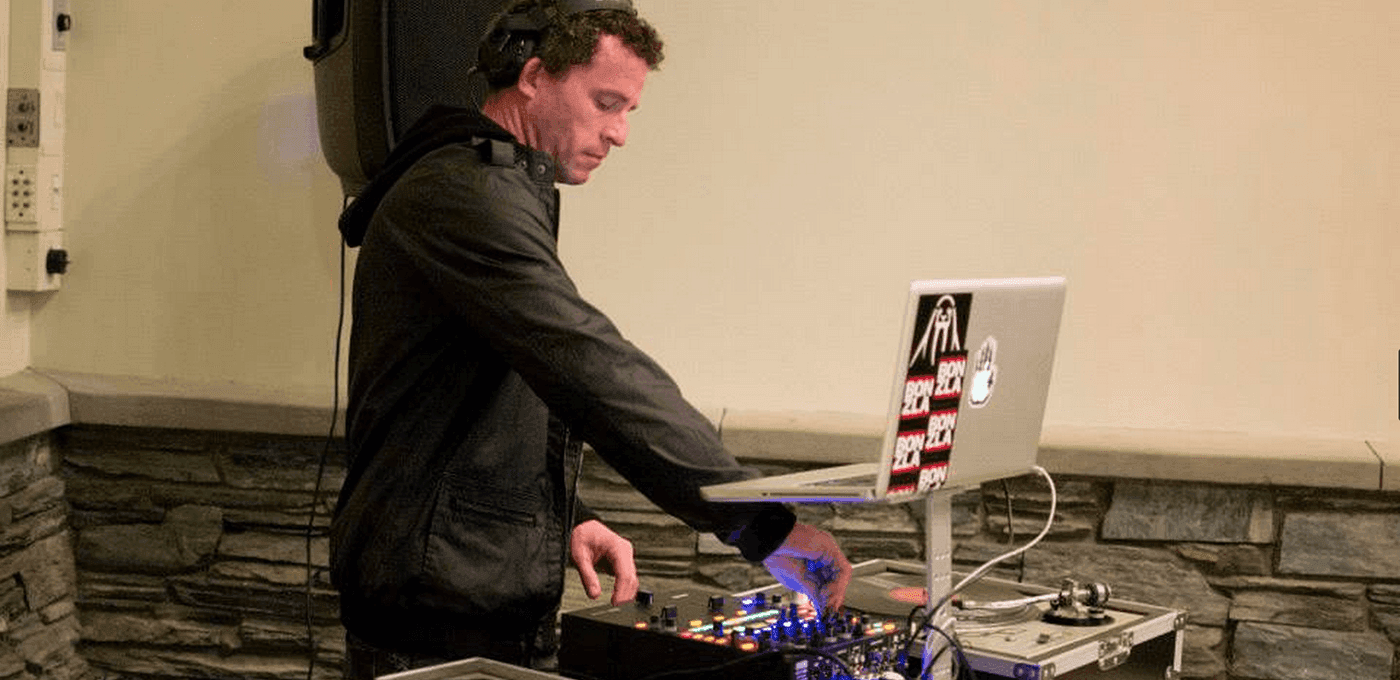 DJ wearing a black jacket and headphones adjusts a mixer beside a laptop on a stand, with a turntable nearby and a stone wall backdrop.