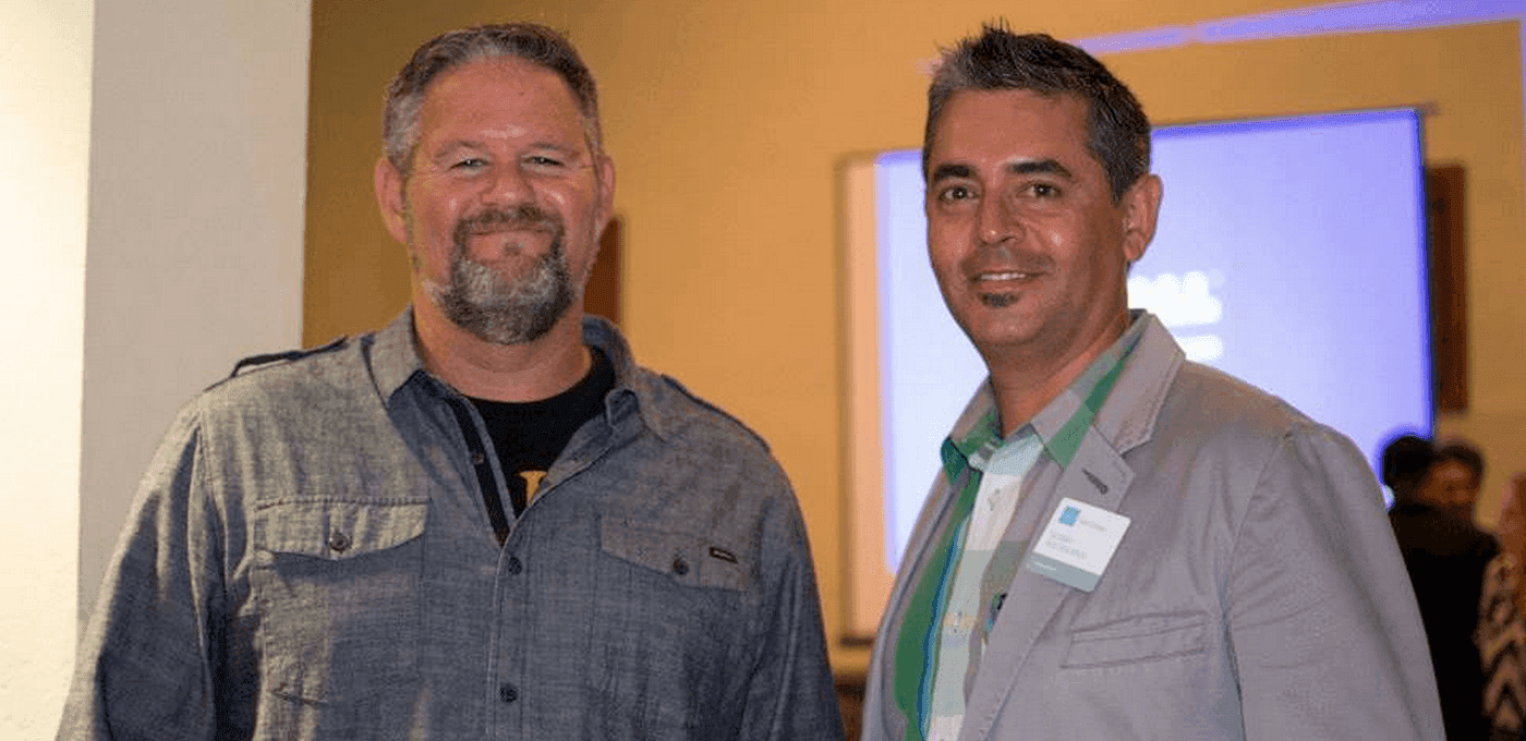 Two people at an indoor event, smiling at the camera; left in a gray denim shirt, right in a light blazer with a name badge, yellow wall behind.
