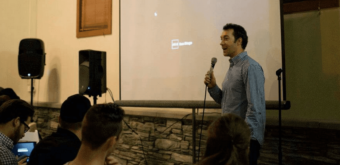A person speaks into a microphone on a small stage in front of a projection screen; audience sits nearby with two large speakers.