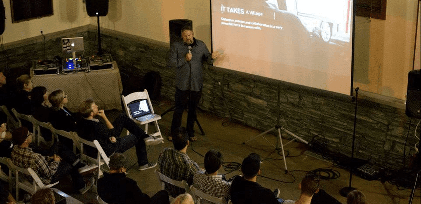 Speaker presenting in a small room, standing by a projected screen; audience sits in white folding chairs, with a laptop and audio equipment on a nearby table.
