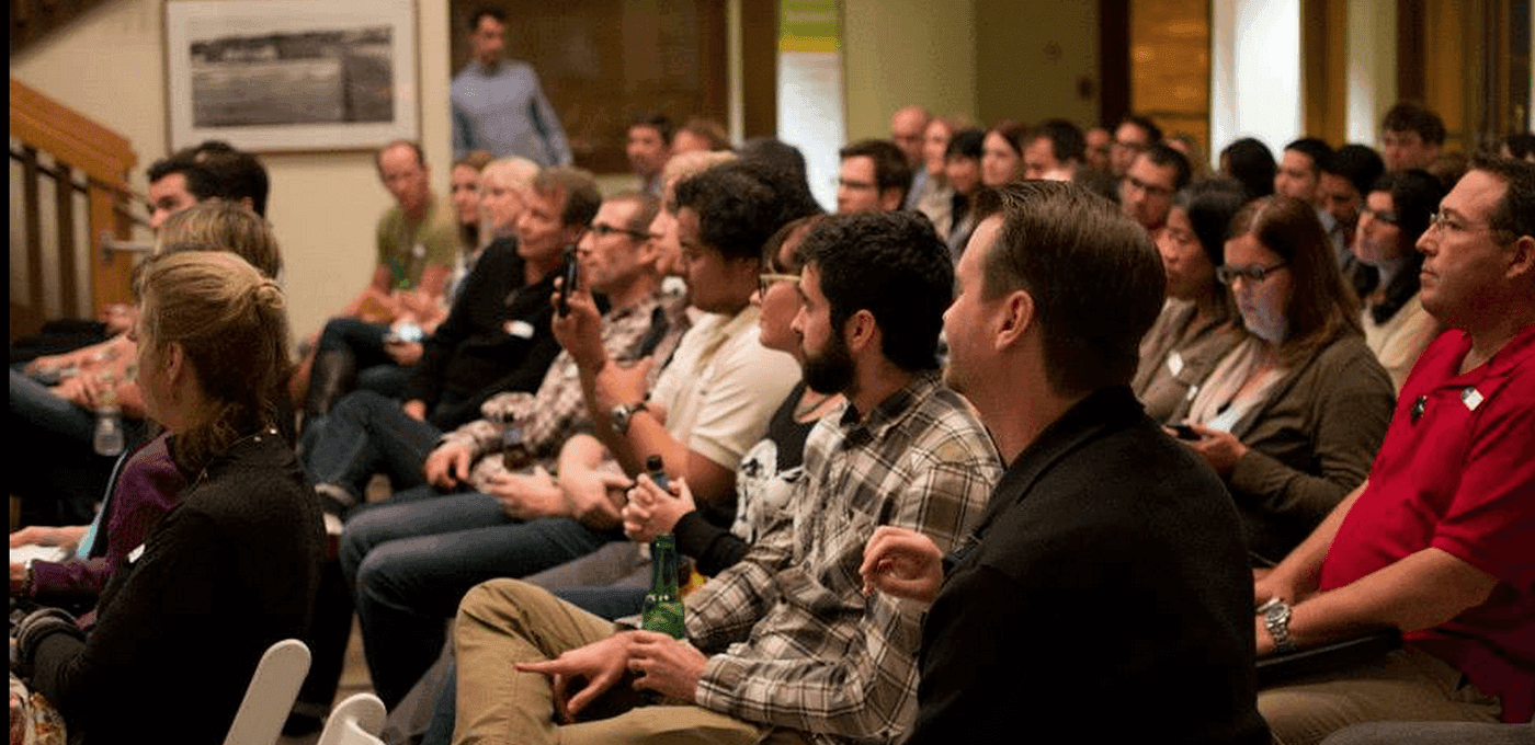 Diverse audience seated in rows at an indoor event, facing forward; some hold drinks, with warm lighting and a few people standing in the back.