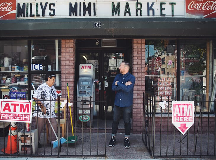 MILLYS MINI MARKET storefront with two people outside; one near an ATM and another with arms crossed in a dark blue shirt, brick facade.