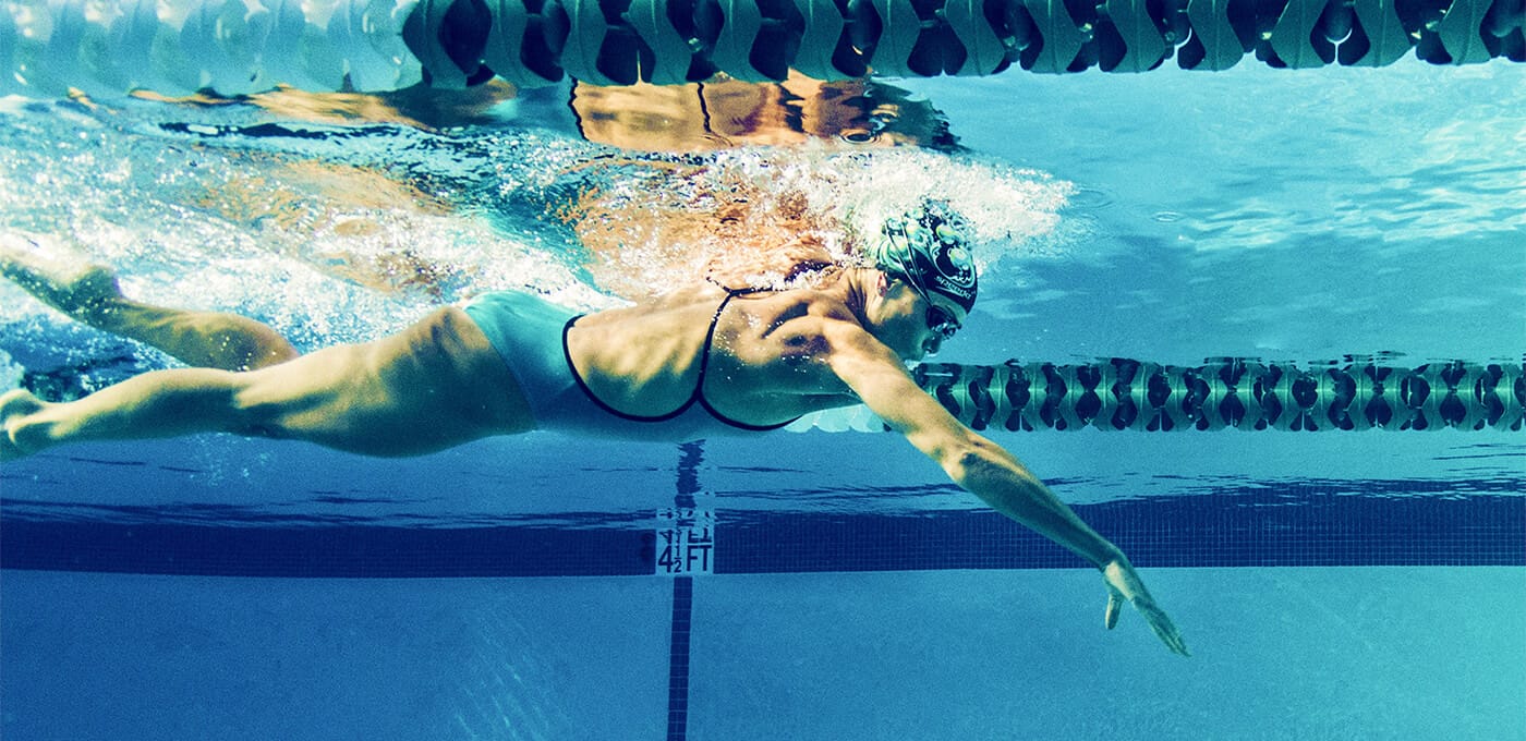 Underwater view of a swimmer in a blue swimsuit, cap, and goggles gliding in a pool lane with one arm extended forward.