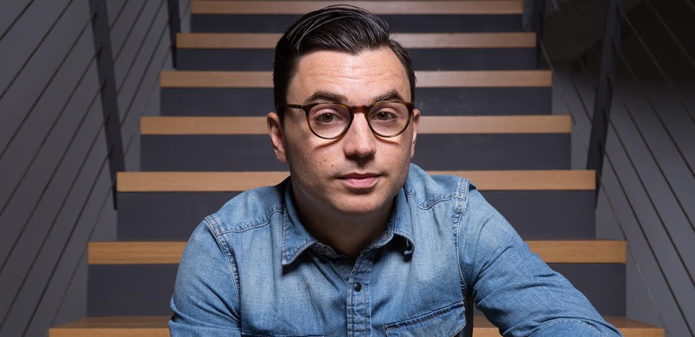 Person with short dark hair and round glasses wearing a blue denim shirt sits on a modern staircase, facing the camera with a neutral expression.
