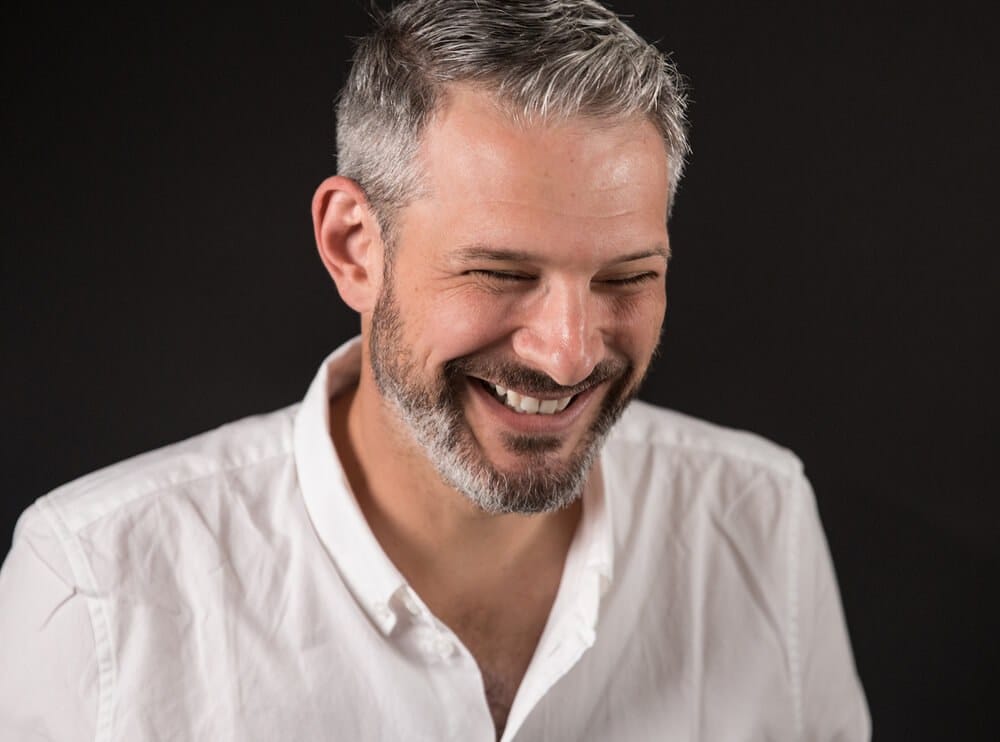 A smiling person with short gray hair and a trimmed beard, wearing a white button-down shirt against a dark background.
