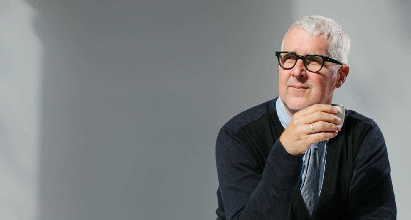 A person with short gray hair and black glasses, wearing a dark cardigan over a light shirt, holds a small cup near their lips, gazing to the right.