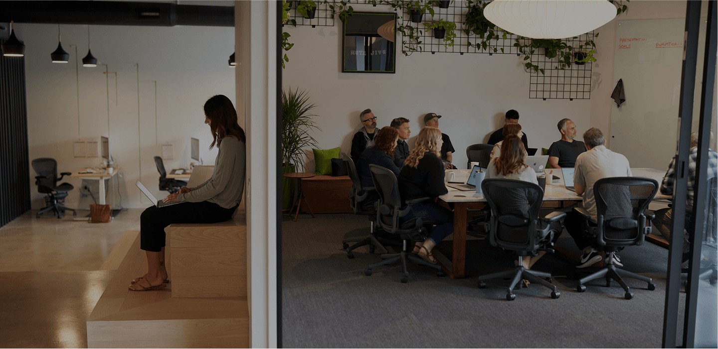 Person sits on a wooden stepped bench with a laptop in an open-plan office; a doorway leads to a conference room with a long table and people at laptops.