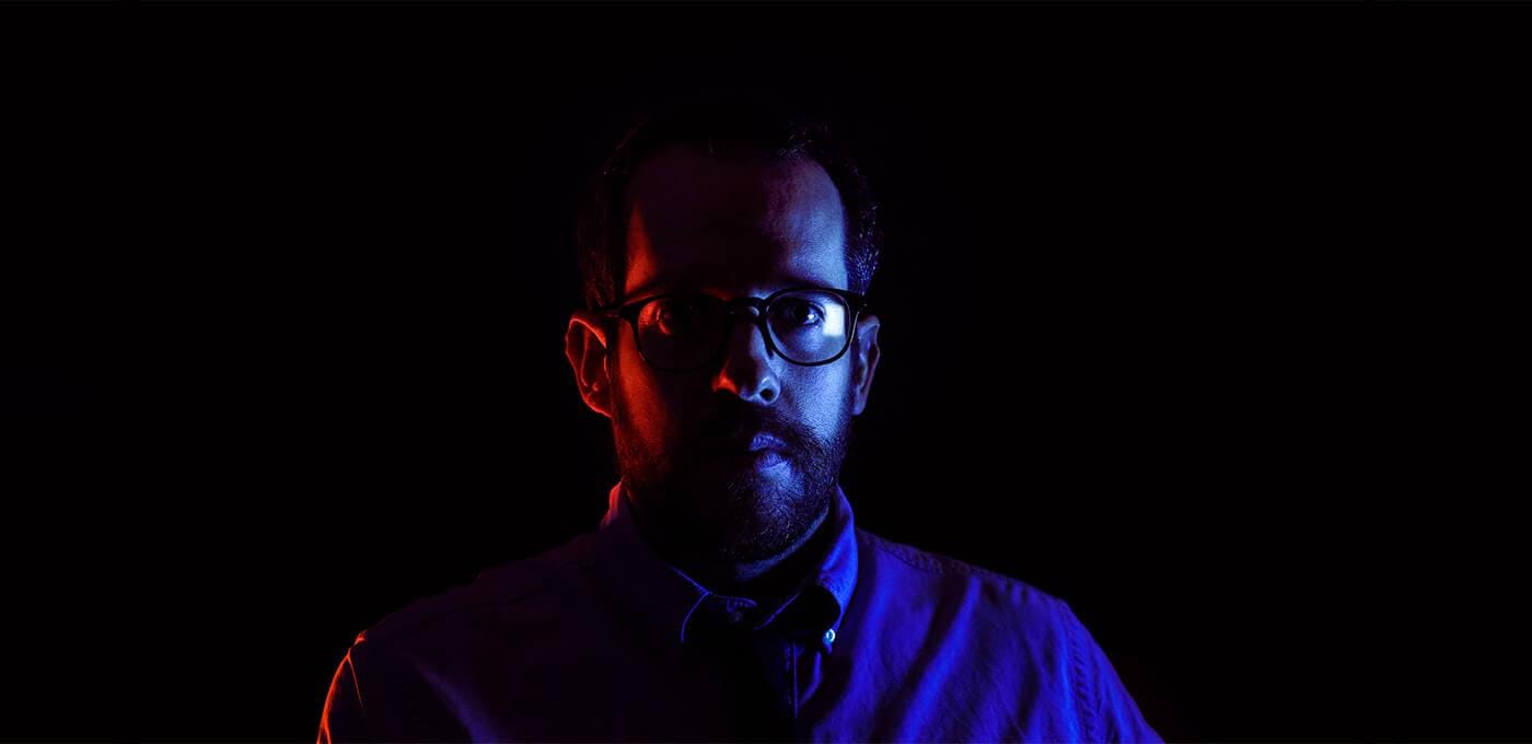 Portrait of a person wearing glasses in a dark setting, lit with red on the left and blue on the right.