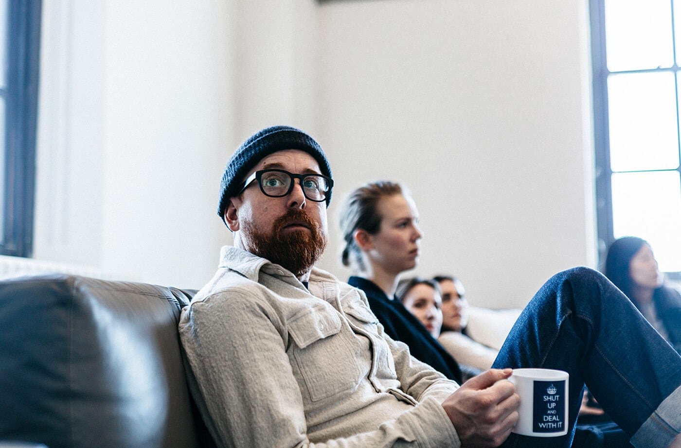 A person with a beard, glasses, and a dark knit cap sits on a couch holding a mug; others in the background near a large window.