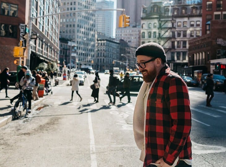 A person in a red-and-black plaid shirt, sweater, black beanie, and glasses smiles while walking on a city street with pedestrians and buildings.