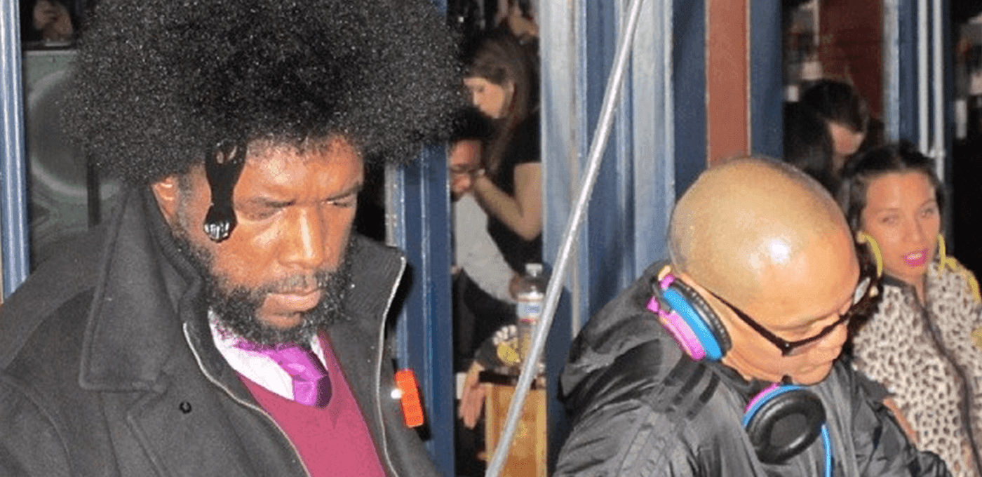 Two people in a busy setting: left with an Afro and beard, pink shirt and dark coat with an orange badge; right bald with colorful headphones and a black jacket.