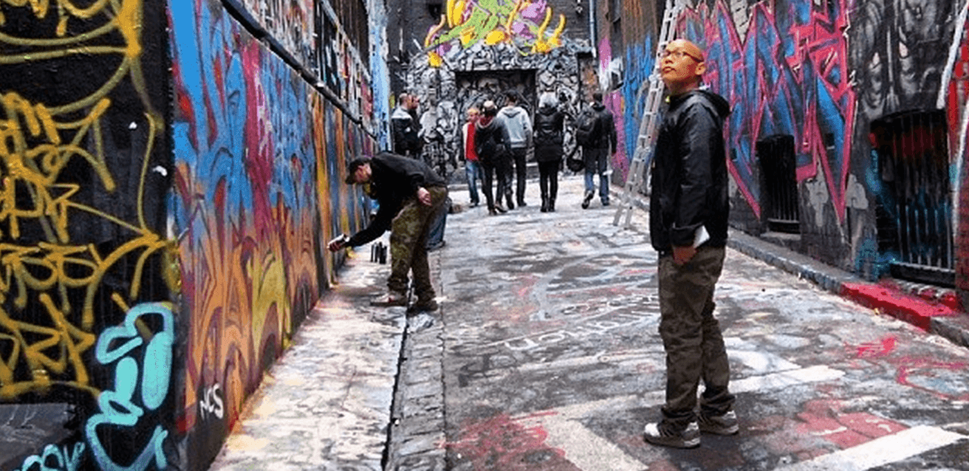 Graffiti-filled alley with colorful murals; a person spray-painting the wall on the left, another stands on the right, and a small group gathers in the background.