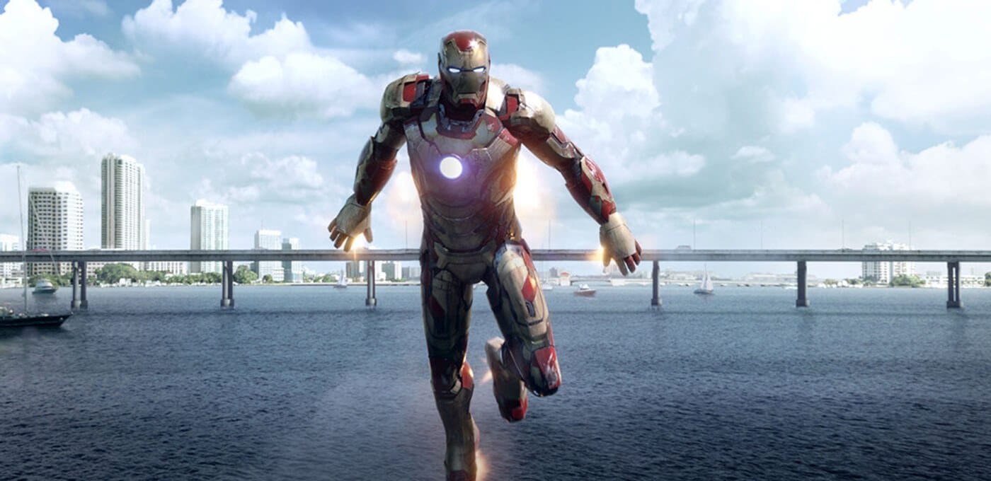 A person in red and gold armor hovers above a wide river, with a city skyline and a bridge in the background beneath a cloudy sky.