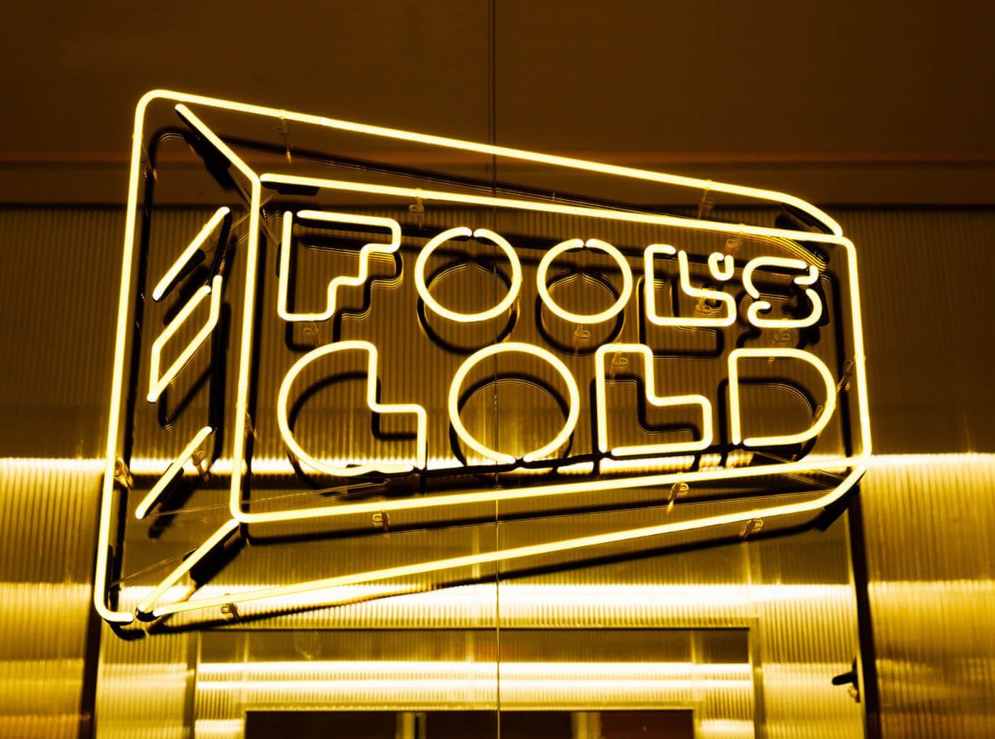 Bright yellow neon sign reading "FOOD GOLD" inside a rectangular frame, mounted on a metallic wall, glowing warmly.