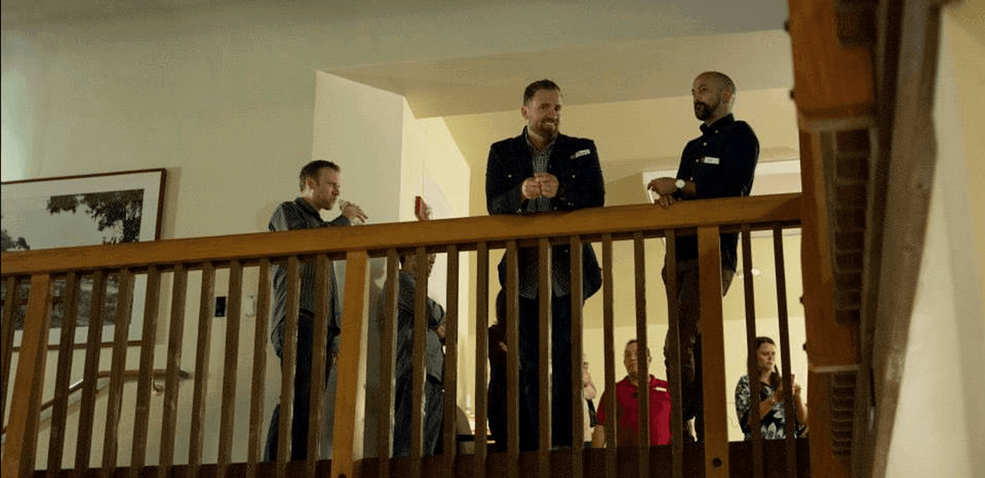 Several people stand behind a wooden railing on an indoor balcony, with one person in a blazer speaking while others listen and clap.