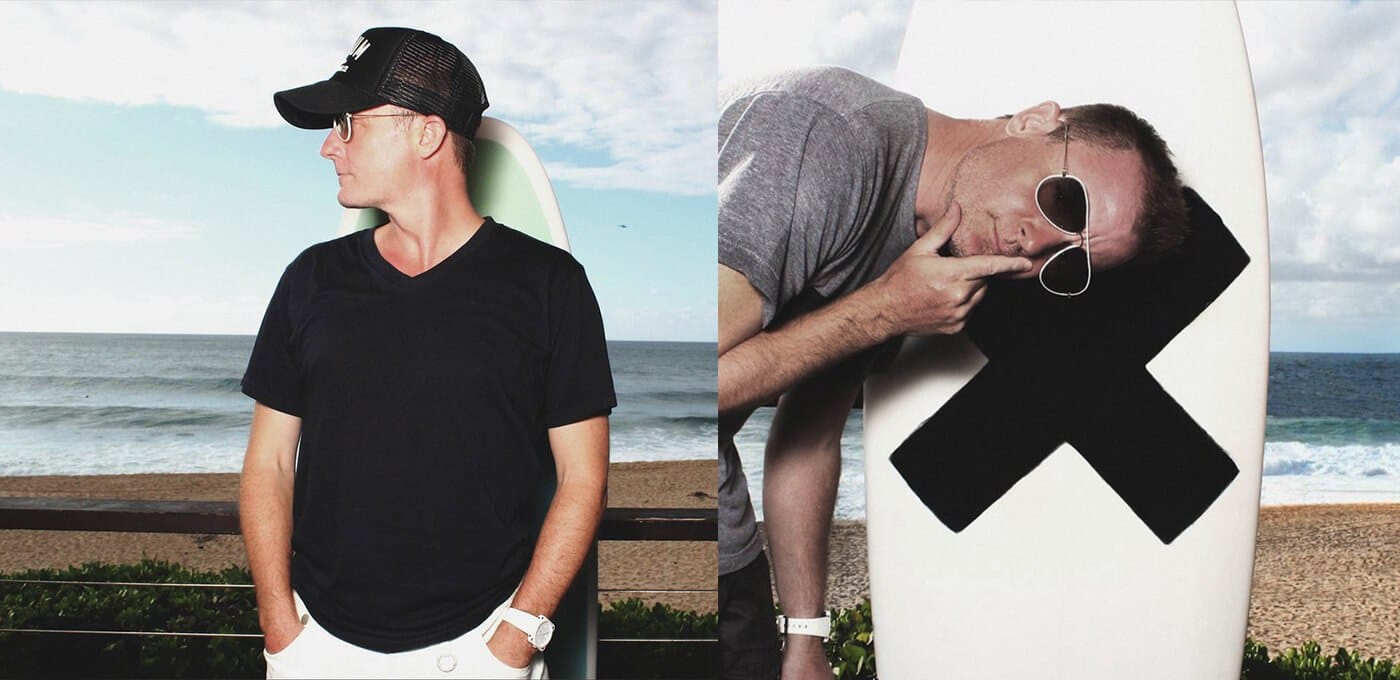 Two people at the beach with surfboards: left in a black T-shirt and cap facing left; right in a gray shirt with sunglasses posing by a white board with a large black X.