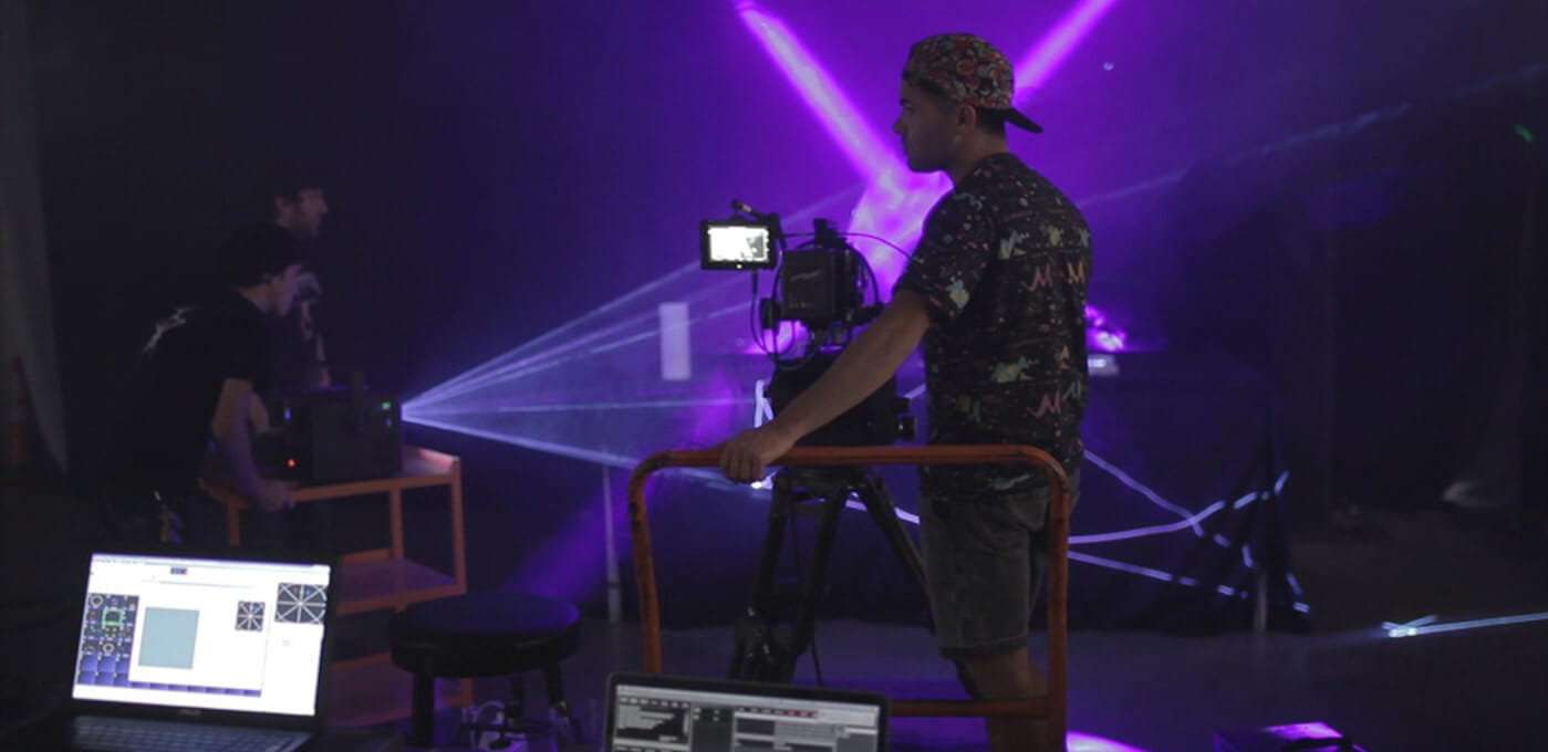 A person in a floral shirt and cap operates a camera on a rolling rig in a dark studio, with purple laser beams and laptops visible in the background.