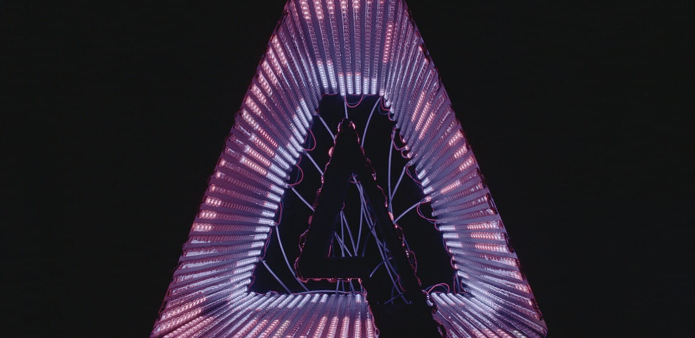 Neon sign in a triangular frame forming the letter A, glowing pink and purple with visible wires against a dark background.
