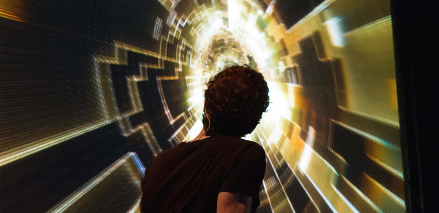 Person with dark hair wearing a black shirt stands in a bright, spiraling tunnel of geometric light panels, viewed from behind.