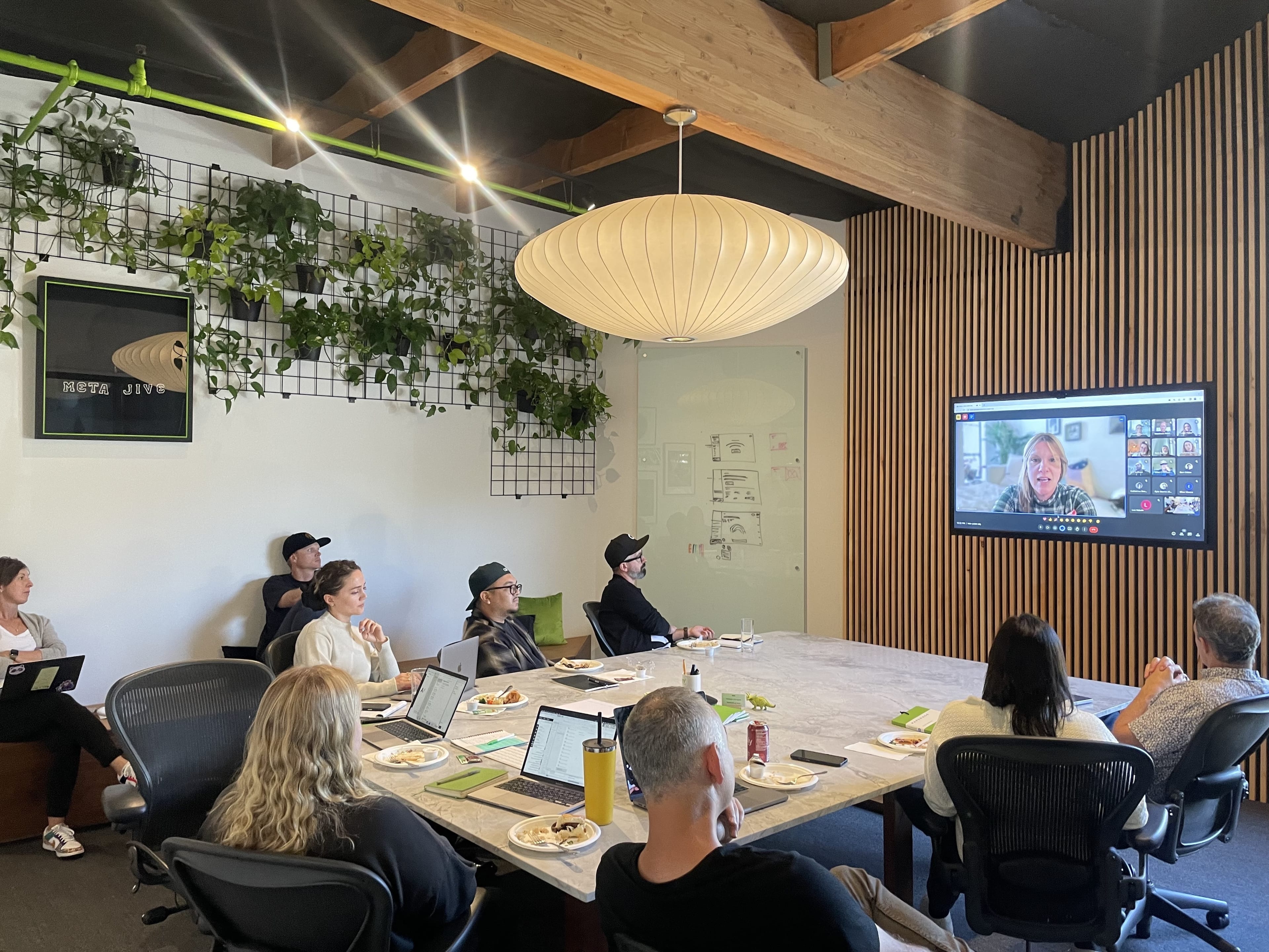 Team in a bright modern meeting room watches a video call on a wall screen; long table with laptops and snacks, plants on a grid wall, and a round pendant lamp.