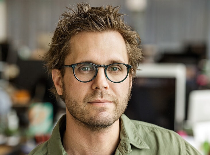 Portrait of a person with short, tousled brown hair and round glasses, wearing a green shirt, in a softly blurred office setting.