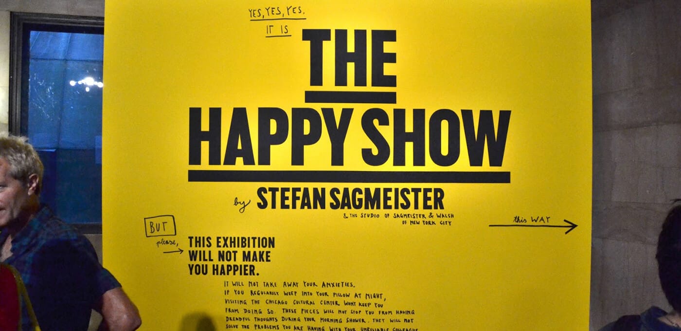 Bright yellow wall with a large bold black poster reading THE HAPPY SHOW by STEFAN SAGMEISTER; partial people visible at the left and right edges.