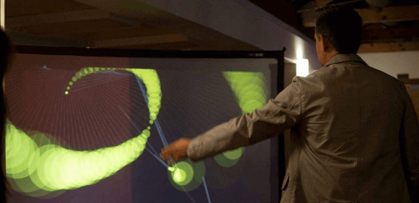 Person in a gray jacket stands with arm extended, pointing at a large interactive screen showing neon green spiral shapes and circles.