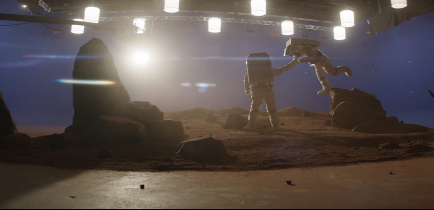 Two astronauts in bulky suits on a rocky Mars-like surface, one reaching to assist the other near a boulder under bright studio lights.