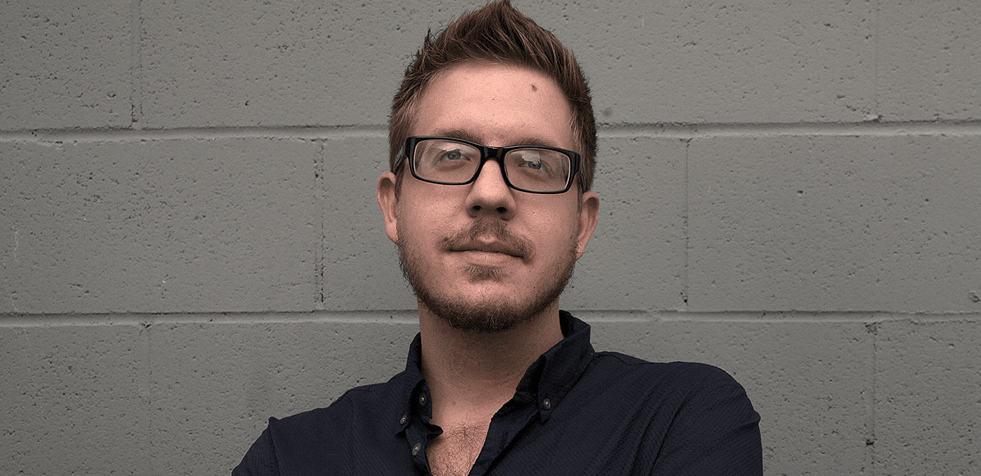 Person with short brown hair and glasses, light stubble, in a dark button-up shirt, standing against a gray cinder block wall, looking at the camera.