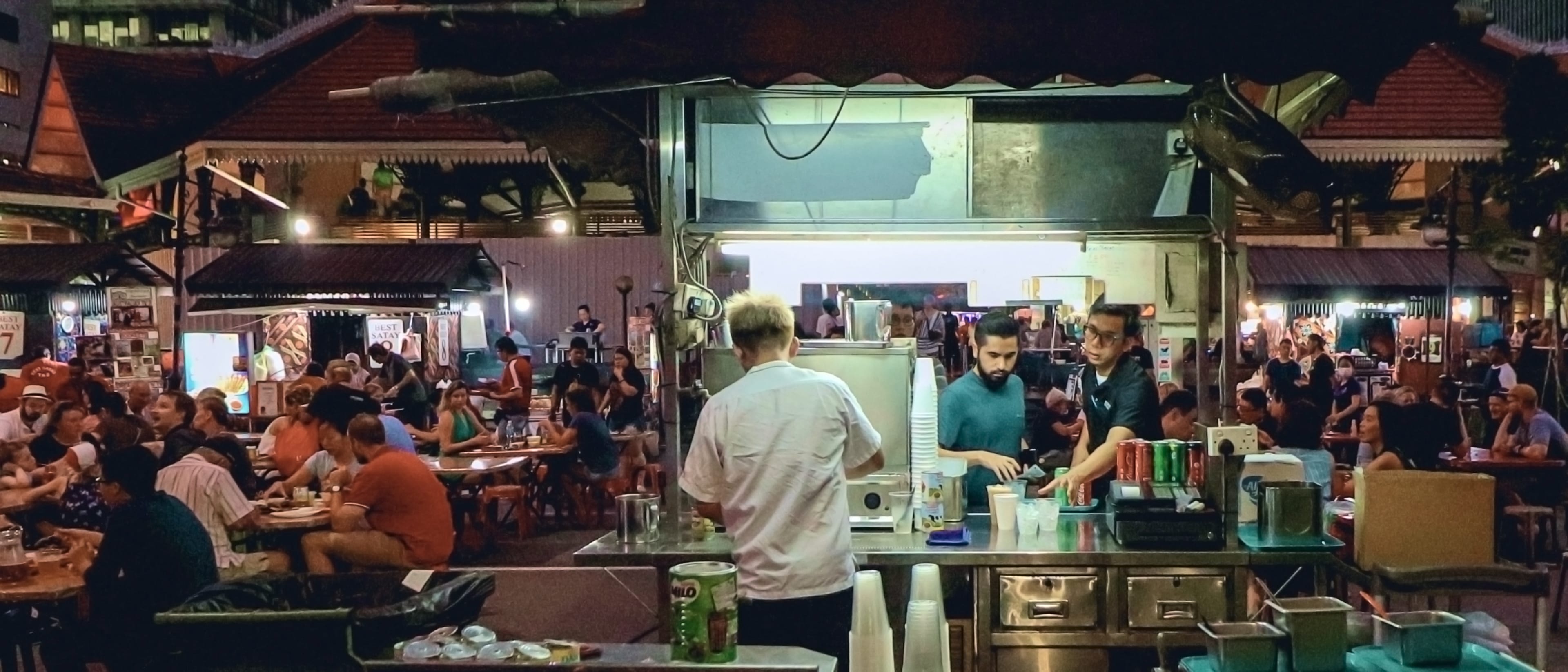 Night market scene with a busy open-air food stall in the foreground; diners fill tables as two staff prepare drinks behind a stainless counter.
